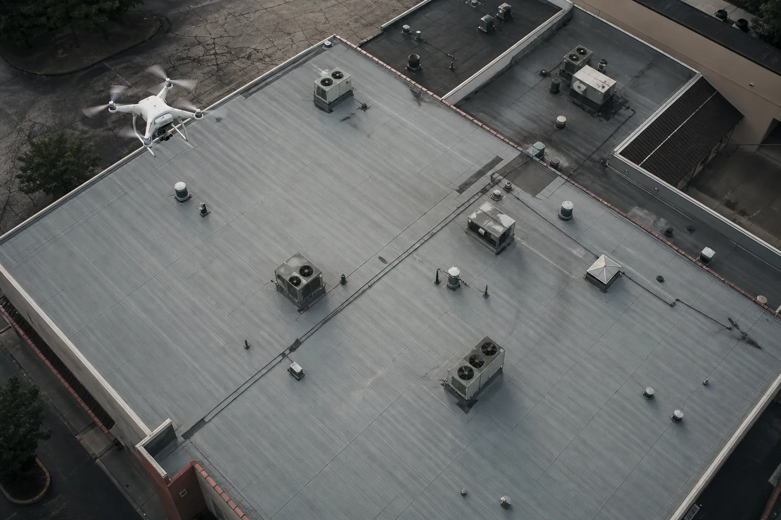 A flat commercial roof with a membrane surface, seams and rooftop HVAC penetrations, photographed from above with a drone visible mid-flight over one corner