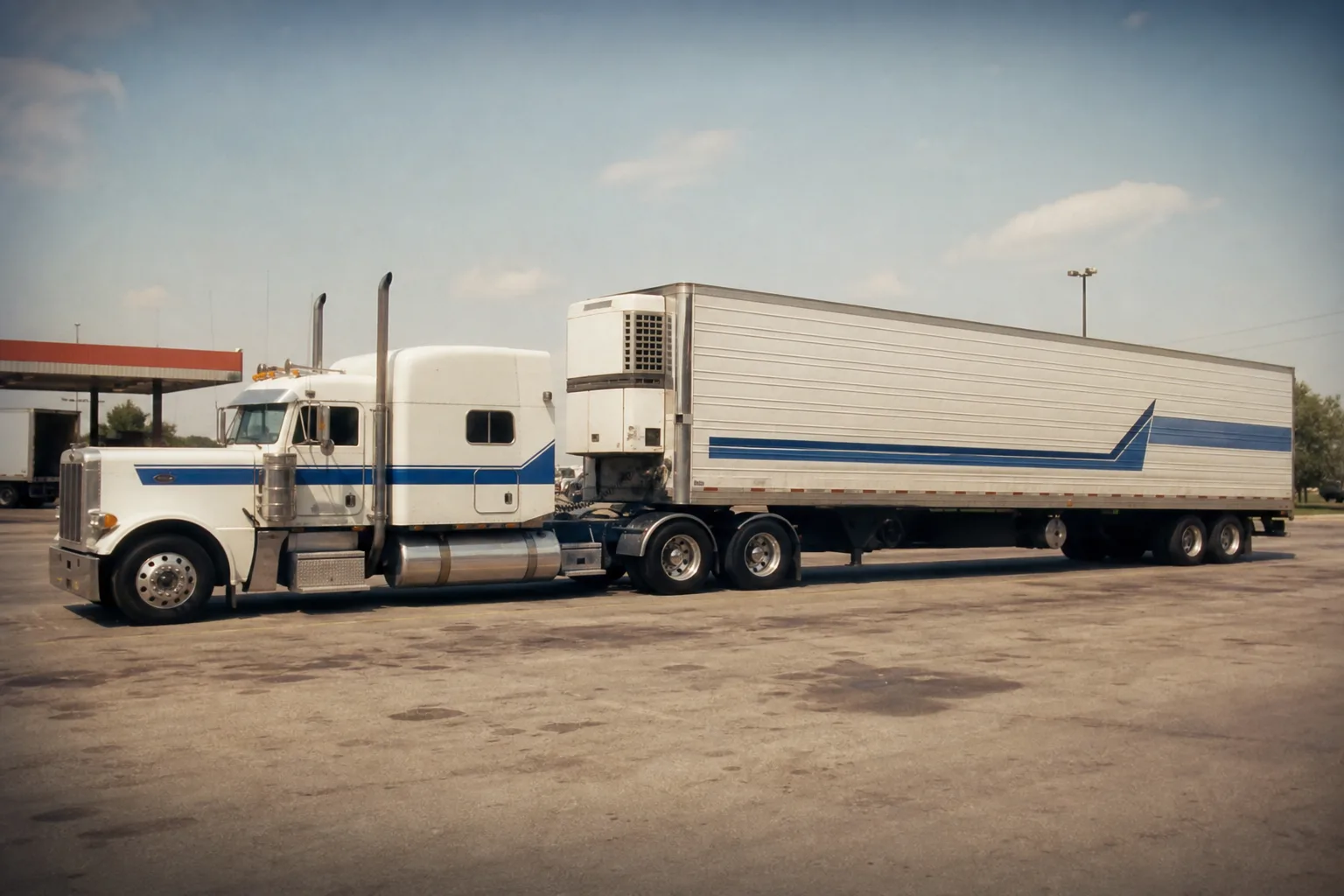 A tractor-trailer parked at a highway truck stop, photographed from the side so the cab door markings and the separate trailer markings are both visible
