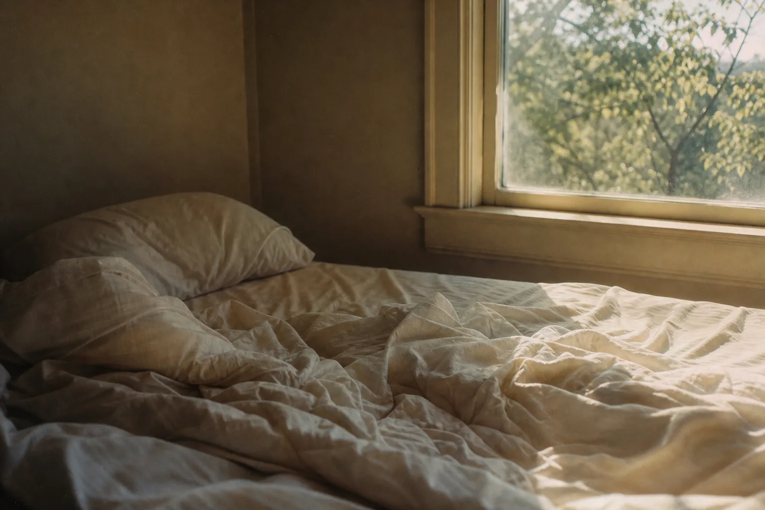 An unmade bed beside a window with early morning light falling across the covers