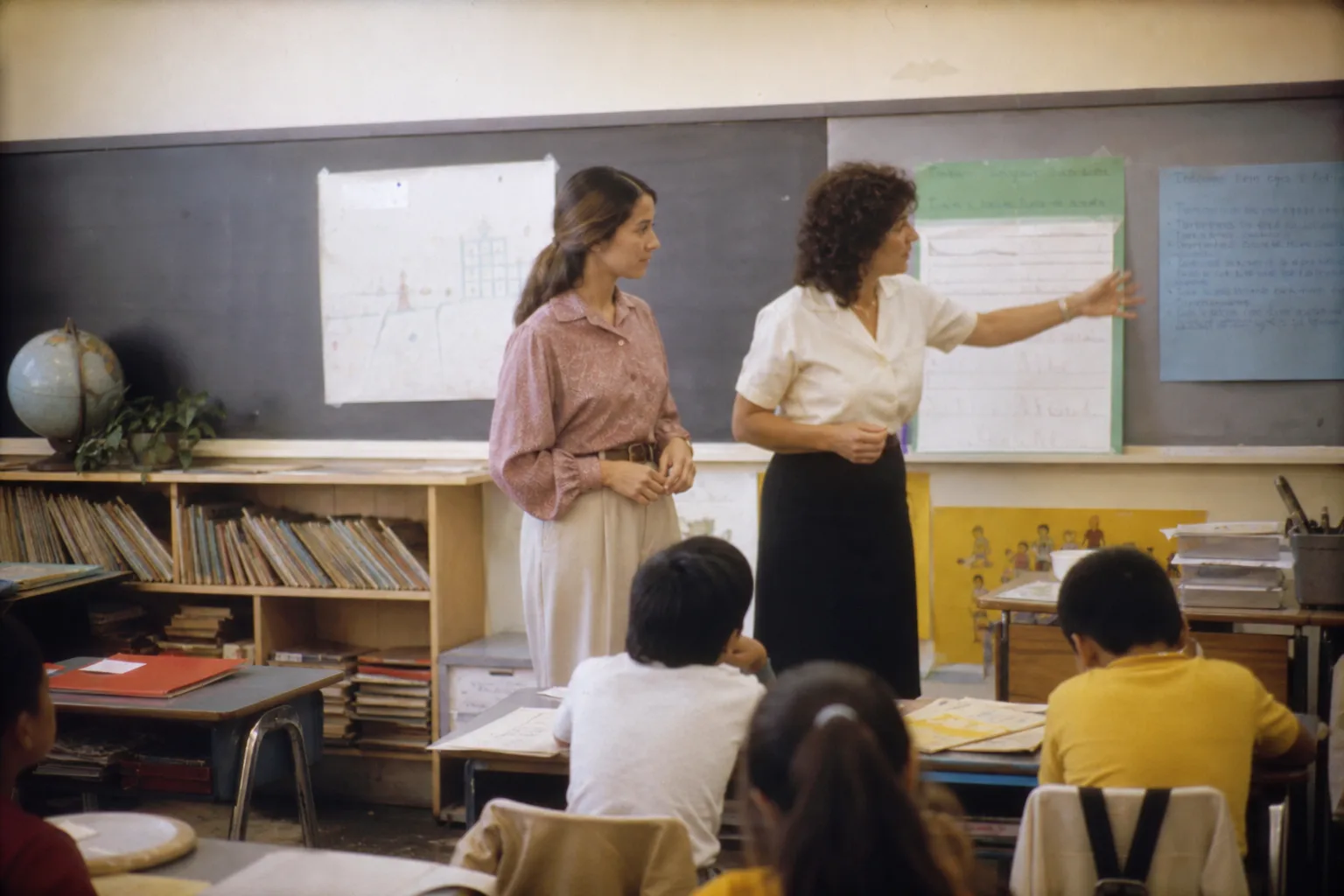 An adult student teacher standing beside a mentor teacher at the front of an elementary classroom in Los Angeles, student desks and chart paper visible