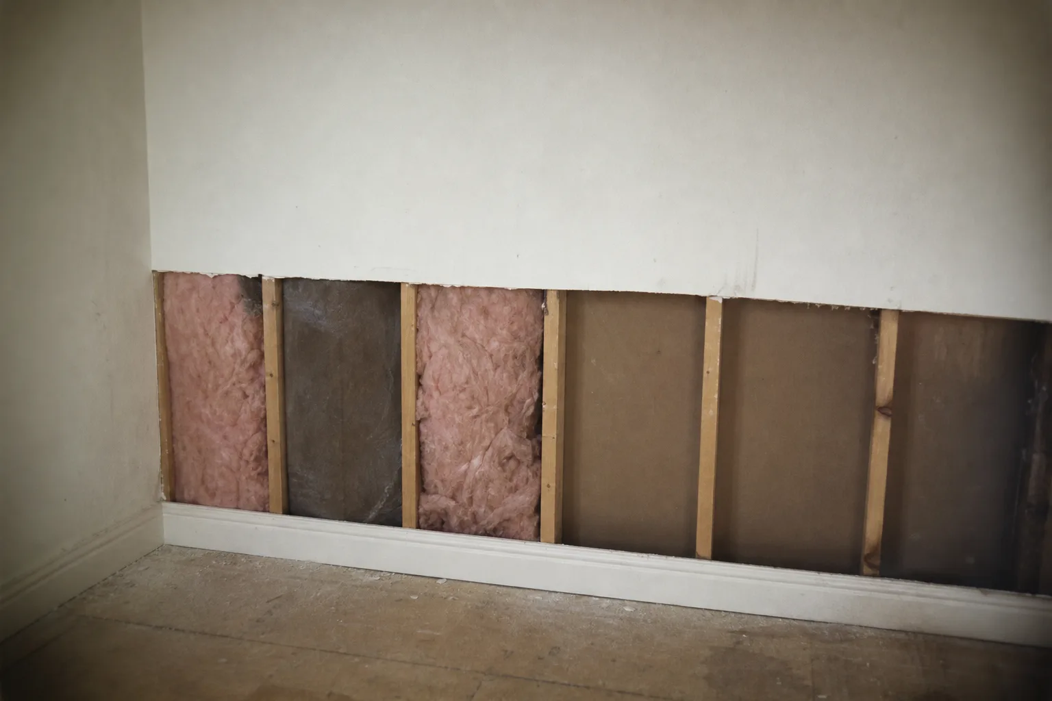 A section of drywall cut away above a baseboard revealing wall studs and insulation in a bare room